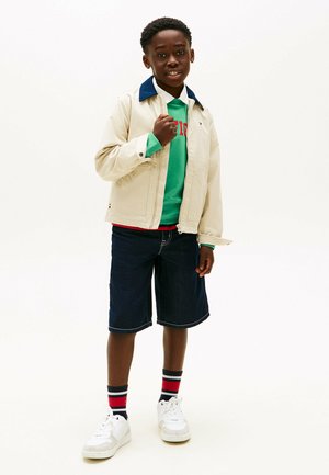 Dreng står, iført beige jakke over grøn sweatshirt, mørke denimshorts, stribede sokker og hvide sneakers på ensartet hvid baggrund.