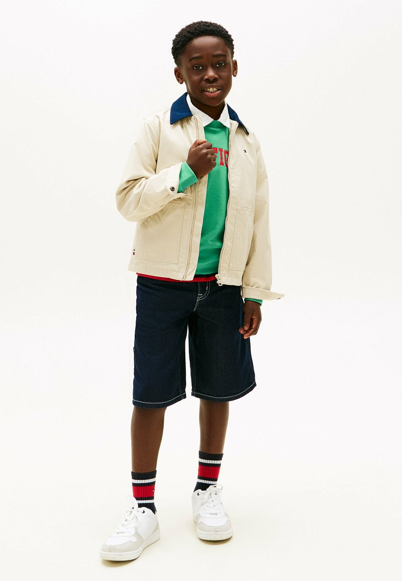 Dreng står, iført beige jakke over grøn sweatshirt, mørke denimshorts, stribede sokker og hvide sneakers på ensartet hvid baggrund.