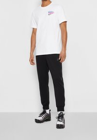 Vit Nike t-shirt med logotyp på framsidan, svarta träningsbyxor med sidostripes, och silverfärgade sportskor med svarta snören och gröna detaljer.