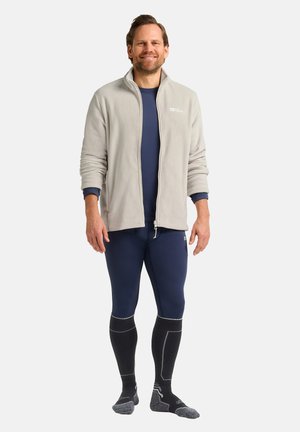 Lichte grijze fleeceselector voor heren over een donkerblauwe lange mouwen top, gecombineerd met marineblauwe leggings en donkere sokken met grijze accenten. Staande pose.