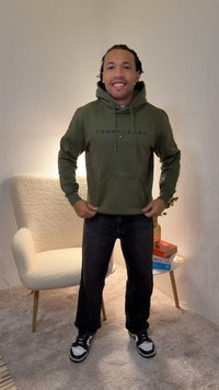Grüner Kapuzenpullover mit "TOMMY JEANS"-Logo, Kängurutasche und Kordelzugkapuze, kombiniert mit schwarzen Hosen und schwarz-weißen Sneakern.