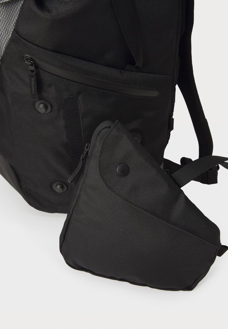Sac à dos noir en tissu avec plusieurs poches zippées et une petite pochette triangulaire. Surface texturée et boutons-pression visibles.