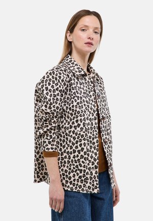 Femme à la peau claire et aux cheveux raides portant une veste boutonnée beige à imprimé léopard noir par-dessus un haut marron et un jean bleu, debout devant un fond blanc.