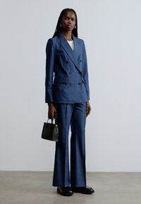 Tamsiai mėlynas dvieilis švarkas su derančiomis platėjamos priekyje kelnėmis, derinamas su maža juoda rankine ir juodais batais. Lygus audinys ir priderintas siluetas.