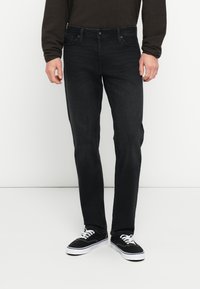 Crne traperice uskog kroja s klasičnim dizajnom s pet džepova, suptilnim šavovima i glatkom teksturom, uparene s crnim tenisicama.