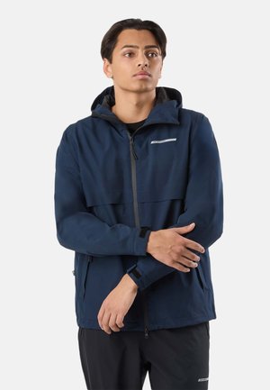 Jeune homme portant une veste à capuche bleu marine zippée et un pantalon noir, ajustant sa manche, debout devant un fond clair uni.