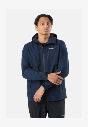 Junger Mann, der eine marineblaue Kapuzenjacke mit Reißverschluss und schwarze Hose trägt, den Ärmel anpasst, vor einem schlichten hellen Hintergrund stehend.