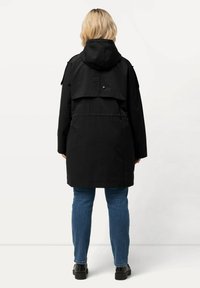 Musta vedenpitävä parka, jossa on huppu, kerroksellinen selkäosa, pitkät hihat, sivutaskut ja rento istuvuus, yhdistettynä sinisiin farkkuihin.