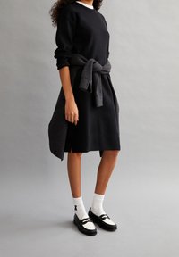 Robe sweat-shirt noire à manches longues et col montant, cintrée par un pull gris foncé. Portée avec des chaussettes blanches et des mocassins noirs.