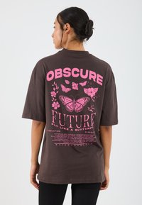 Brown oversized T-shirt featuring large pink graphic text and butterfly designs on the back. Short sleeves, relaxed fit, cotton material.