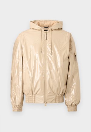 Beige glanzende gewatteerde jas met capuchon, voorrits, trekkoorden, elastische manchetten en een ritszak op de linker mouw, getoond tegen een effen achtergrond.