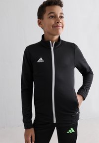 Schwarze Sportjacke mit weißem Reißverschluss, mit einem Adidas-Logo auf der linken Brust. Entworfen mit Stehkragen und flachen Bündchen.