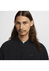 Zwarte Nike-jack met een rits aan de voorkant, hoge kraag en geborduurd logo. Heeft een gladde textuur en een minimalistisch ontwerp.