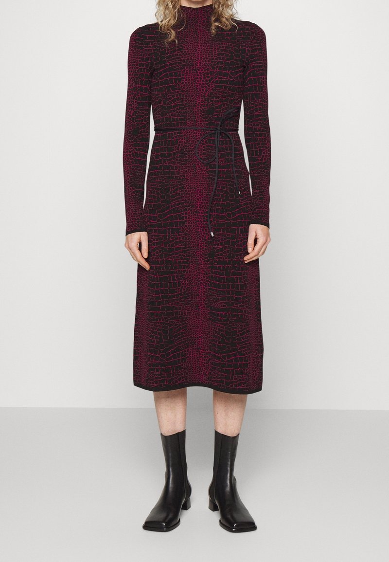 Femme portant une robe midi à manches longues, noire et rouge à motifs, avec une ceinture à la taille, associée à des bottes noires mi-mollet, debout contre un fond blanc.