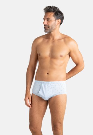 Homme torse nu avec des cheveux courts et foncés et une barbe, portant un slip bleu clair avec de petits motifs d'ancre, debout devant un fond uni clair.
