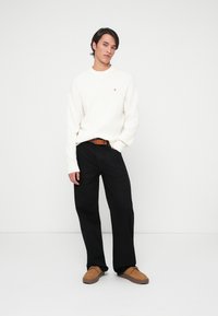 Pulover alb cu striații și un mic logo roșu, pantaloni negri evazați și pantofi maro cu suprafață texturată. Un outfit simplu și casual.