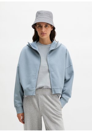 Femme portant un sweat à capuche zippé bleu clair, un t-shirt assorti, un pantalon de survêtement gris, et un chapeau bob gris clair avec logo sur un fond uni.