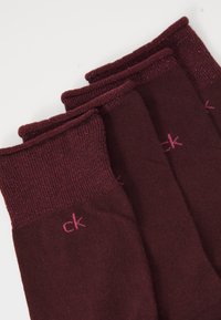 Quattro paia di calzini di un intenso bordeaux scuro con polsini rigati scintillanti e piccoli loghi "ck" rosa vicino al bordo superiore su uno sfondo bianco.