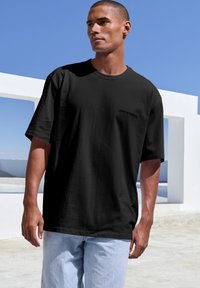 Zwarte oversized t-shirt met korte mouwen en een ronde hals, met een subtiele "CPH STUDIOS" logo aan de voorkant, gecombineerd met lichtblauwe jeans.