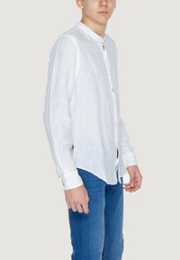 Camicia bianca in lino con colletto alla coreana, maniche lunghe e chiusura con bottoni. Abbinata a jeans blu, per un look casual.
