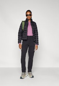 Zwarte gewatteerde jas met hoge kraag, paarse lange mouwen top, zwarte cargobroek, grijze en teal wandelschoenen en groene rugzakriem.