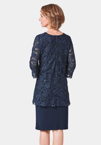 Navyblaues Spitzenoberteil mit floralen Stickereien, durchsichtigen langen Ärmeln und einem einfarbigen navyblauen Rock. Die Rückseite hat einen runden Ausschnitt und einen dezenten Reißverschluss.