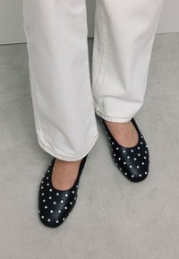 Zwarte leren ballerina's met een ronde neus, versierd met zilveren studs in een patroon, gecombineerd met witte, losse broek.