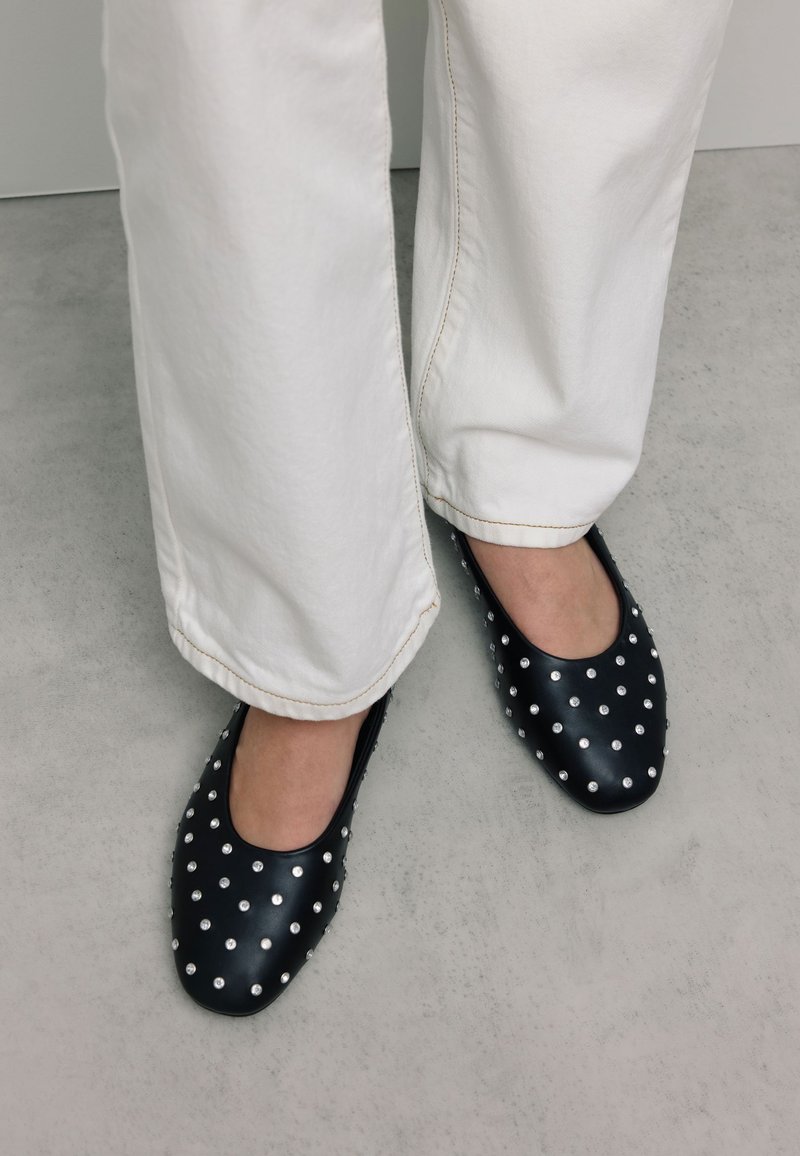 Zwarte leren ballerina's met een ronde neus, versierd met zilveren studs in een patroon, gecombineerd met witte, losse broek.