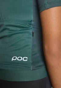 Tricou sportiv de culoare verde închis, cu mâneci scurte, având un mic buzunar cu fermoar și un logo alb "poc" în partea de jos. Textură de țesătură netedă.