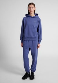 Femme debout portant un sweat à capuche bleu et un pantalon de survêtement assorti avec des baskets noires sur un fond clair uni.