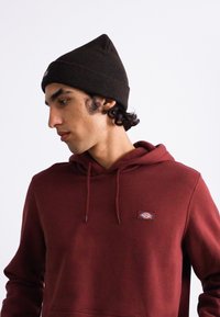 Sweat à capuche rouge avec des manches longues, en tissu texturé et avec un cordon de serrage. Porté avec un bonnet côtelé marron foncé, présentant un profil rapproché.