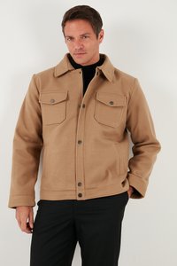 Hombre vestido con una chaqueta de lana color marrón claro con botones y dos bolsillos en el pecho, sobre un cuello alto negro y pantalones negros, de pie frente a un fondo blanco.