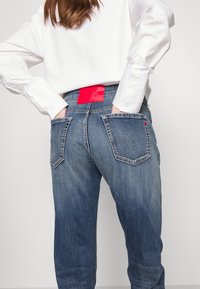 Jeans in denim blu con una finitura sbiadita, dotati di due tasche posteriori e un'etichetta rossa in vita. Abbinati a una camicia bianca a maniche lunghe.