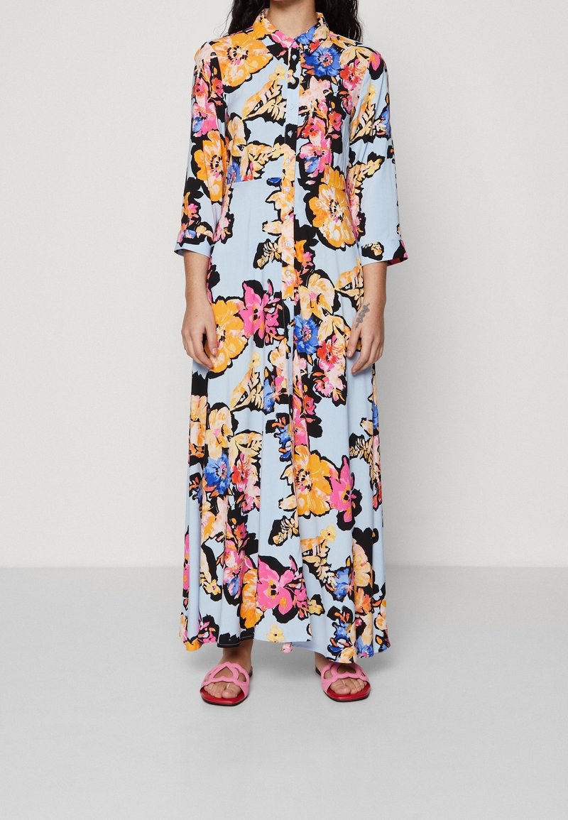 Personne portant une longue robe bleue claire boutonnée avec de grands imprimés floraux jaunes, roses et bleus et des sandales roses, debout sur un sol gris.
