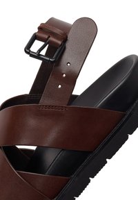 Bruine leren sandaal met zwarte verstelbare gesp en dikke zwarte zool, schuine aanzicht die de voor- en zijdetails toont.