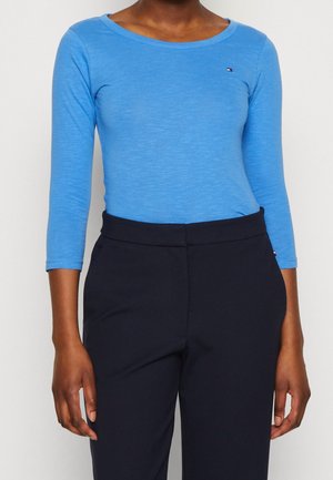 Kobieta ubrana w dopasowaną, niebieską bluzkę z długim rękawem i czarne spodnie z wysokim stanem, stojąca na tle jednolitego, białego tła.