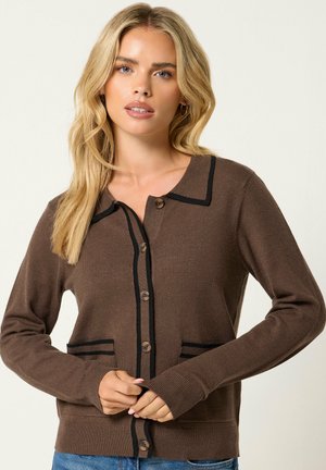 Donna bionda che indossa un cardigan marrone abbottonato con bordi neri e jeans blu, in piedi davanti a uno sfondo chiaro e semplice.