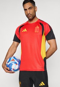 Raudonas ir juodas trumpomis rankovėmis džersis su tekstūruotu dizainu, geltonomis detalėmis ir Belgijos emblemą. Žaidėjas laiko raštuotą futbolo kamuolį.