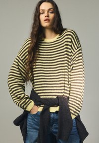 Pruhovaný sveter v žltých a hnedých odtieňoch s voľným strihom a rebrovanými manžetami, kombinovaný s modrými džínsami a tmavým svetrom zviazaným v páse.