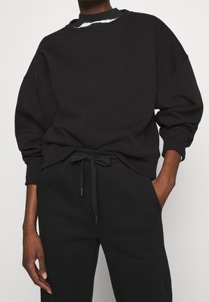 Osoba ubrana w czarną, oversize'ową bluzę i czarne spodnie dresowe ze ściągaczem, jedna ręka w kieszeni, druga swobodnie zwisa przy boku.