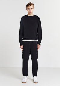 Felpa nera con colletto a girocollo e finitura testurizzata abbinata a pantaloni cargo neri con tasche laterali. Indossata con scarpe da ginnastica bianche.