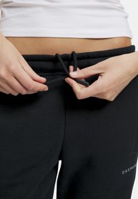 Sorte joggebukser med elastisk midje og snøring, med en glatt tekstur og en liten logo på venstre side.