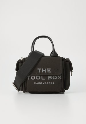 Musta kangaskotelo, jossa on nahkaosuuksia, suorakulmainen muoto, ylhäällä kahvat, irrotettava hihna, jossa on valkoinen teksti "THE TOOL BOX" ja "MARC JACOBS."