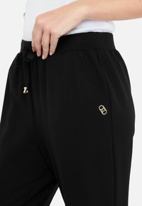 Pantaloni jogger neri realizzati in tessuto liscio, con vita regolabile tramite cordino e dettagli in hardware color oro sul lato.