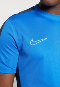 Blå idrottströja med korta ärmar, rund halsringning och en kontrasterande marinblå detalj på axeln. Broderad vit Nike-logga.