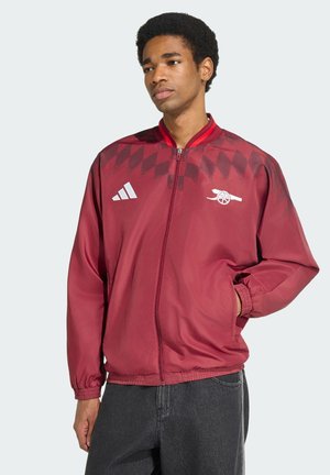 Junger Mann trägt eine rote Jacke mit Reißverschluss mit Adidas- und Arsenal-Logos, steht mit einer Hand in der Tasche vor einfachem Hintergrund.