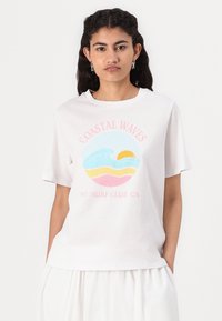 Balta kokvilnas T-krekls ar krāsainu grafiku, kurā attēlotas viļņi un saule zilā, dzeltenā un rozā krāsā, ar tekstu "COASTAL WAVES 97 SURF CLUB CA".