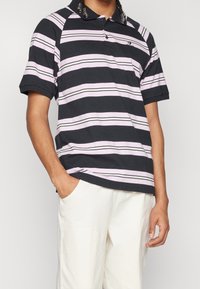 Kortärmad polotröja i svart med rosa och vita horisontella ränder, med krage, två knappar och en subtil logo på bröstet.