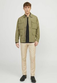 Olivgrön, lättviktsjacka med fyra framfickor, svart polo under, beige slim-fit byxor och svarta skor. Slät textur.