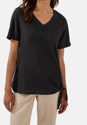 Kvinde iført en sort V-hals kortærmet top med en detalje i form af knude i siden, kombineret med lysebeige bukser mod en ensfarvet baggrund.